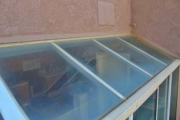 Before: A glass roof installed at an angle against a stucco exterior wall, revealing the interior of a room beneath it through dirty and hard water stained glass showing the need for professional window cleaning.