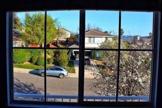 After: View through a freshly cleaned window showing a suburban street with parked cars, green trees, and a house with a driveway. A perfect view delivered after expert window cleaning service.
