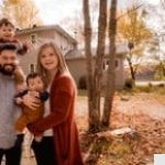 A smiling family of four poses outdoors in front of a house with autumn leaves on the ground. The father holds a toddler on his shoulders and the mother holds a baby, all dressed warmly.