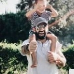 A bearded man wearing sunglasses gives a thumbs up while carrying a smiling child on his shoulders outdoors, with greenery in the background.