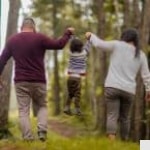 A couple walks through a forest, each holding one hand of a small child between them, lifting the child playfully as they walk along a path surrounded by trees.