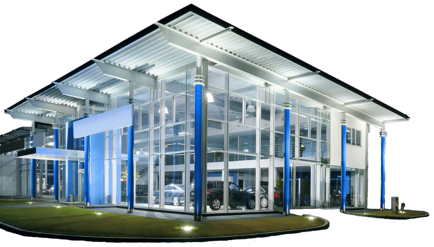 Modern glass-walled car dealership building with bright blue accents, large windows, and a well-lit interior displaying several cars. The sleek structure features a flat overhanging roof, visible metal framework, and benefits from regular commercial power washing.