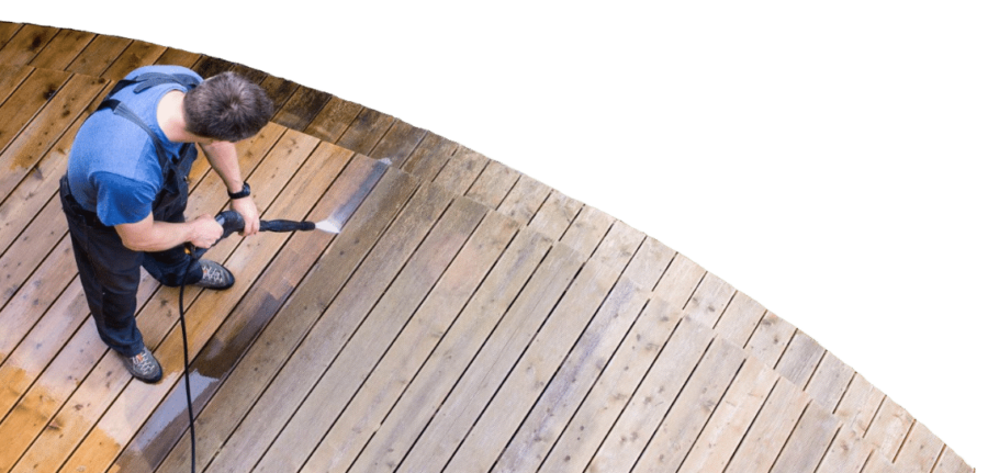 A person uses a pressure washer for deck and fence cleaning, revealing a lighter, cleaner section of the wooden deck compared to the still-dirty area.