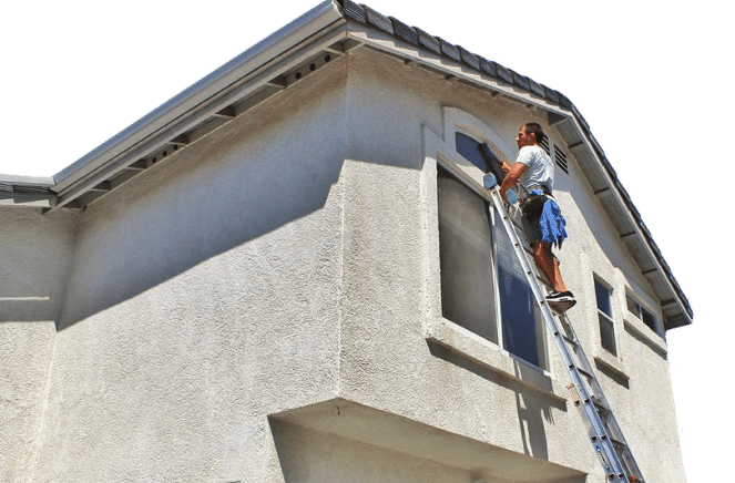 A person standing on a ladder paints the trim around an upper-story window of a light-colored house under bright daylight, after finishing some gutter cleaning.