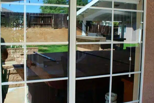 After: A glass window with white grid lines reflects a backyard scene, patio, green grass, and a fence. Thanks to recent window cleaning, a person’s partial reflection and some indoor furniture are also clearly visible in the sparkling glass.