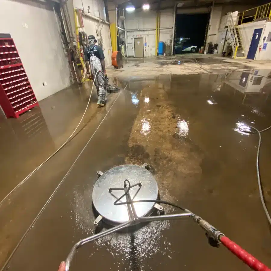 A worker operates a large floor cleaning machine on a wet concrete surface in a spacious industrial area.