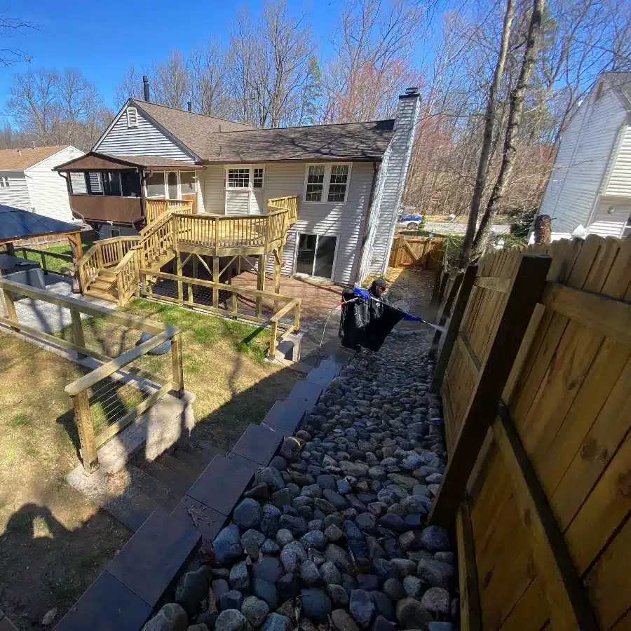Backyard view featuring a wooden deck and a sloped path of stones leading to a fence, under a clear blue sky.