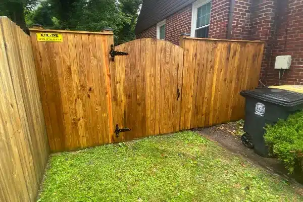 After: Wooden garden gate with a curved top, surrounded by a fence, next to a black trash bin and green grass.