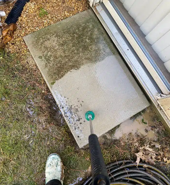 A concrete slab with water puddles, a pressure washer nozzle pointed at the surface, and muddy grass nearby.