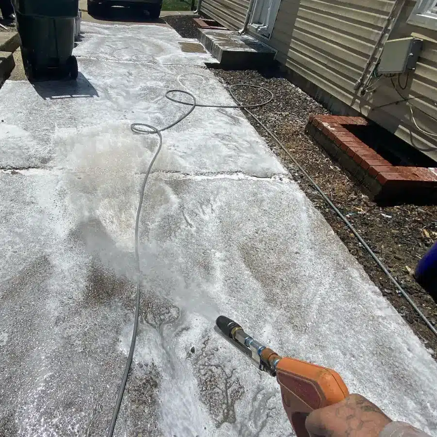 A person holds a pressure washer, spraying water and foam onto a concrete driveway with a trash bin visible nearby.