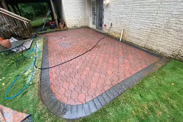 After: A patio with red pavers and a stone border, wet from cleaning, adjacent to a house with a door and garden hose.