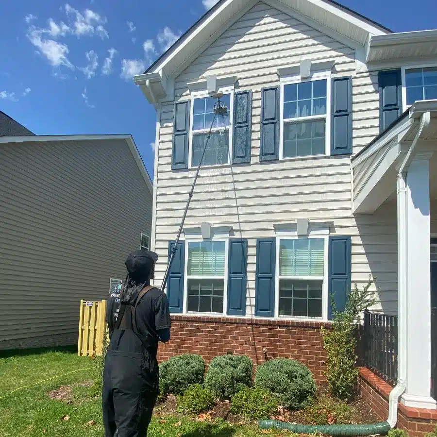 A person in overalls is using a long pole to clean a high window on a two-story house with blue shutters.